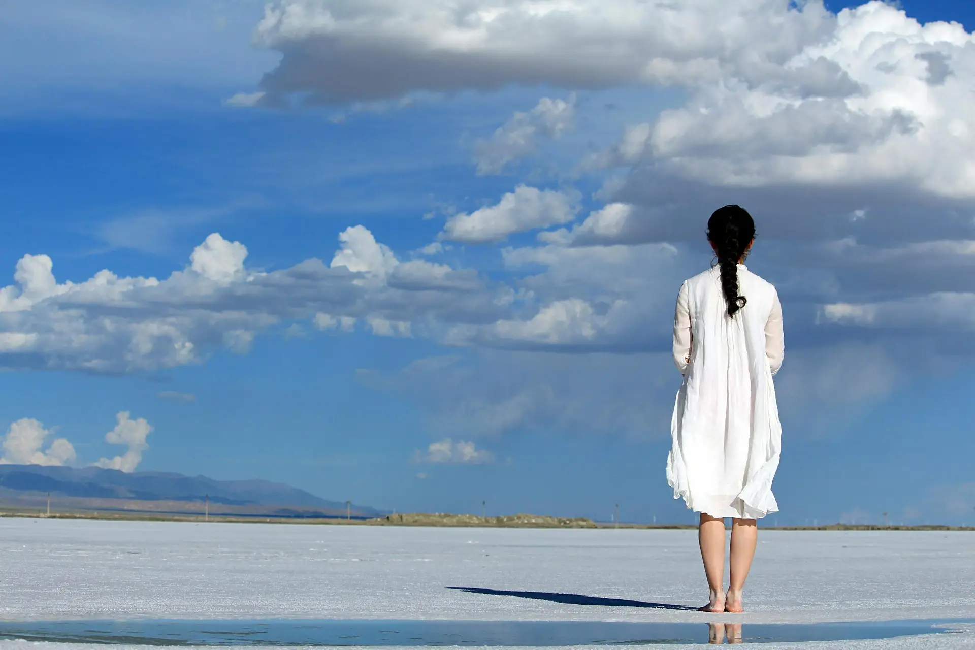 Woman in white dress with braided hair stands barefoot on a vast salt flat under blue sky with clouds.