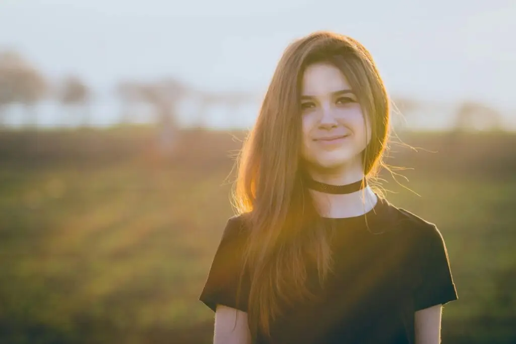 Smiling young woman standing outdoors in a field at sunset, looking hopeful in warm golden light.