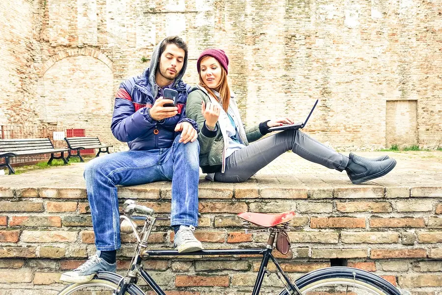 Young man and woman sitting on a brick wall outdoors, looking at a smartphone and laptop, with a bicycle nearby.