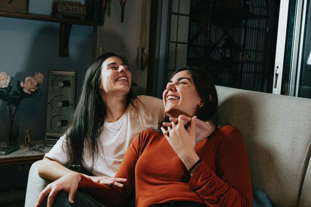 Two women laughing together while sitting close on a couch in a cozy, warmly lit room.