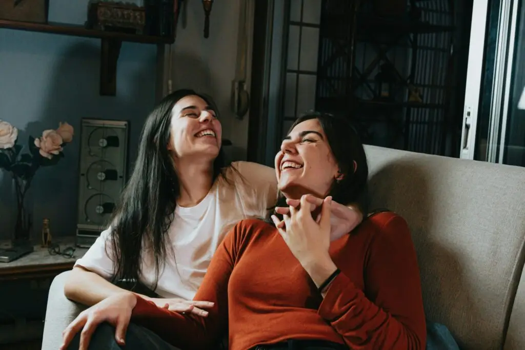 Two women laughing together while sitting close on a couch in a cozy, warmly lit room.