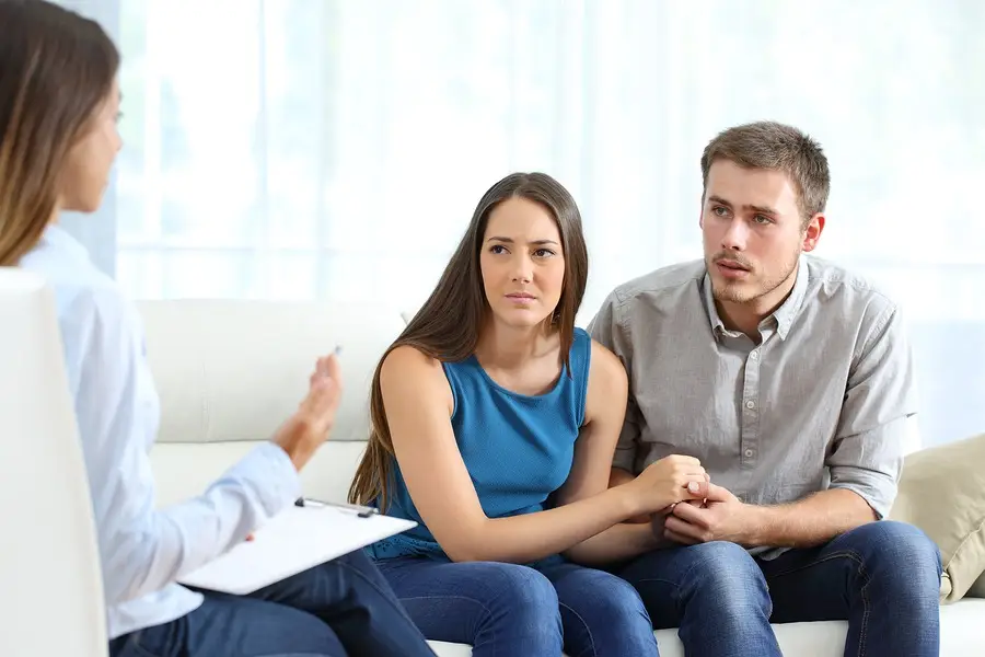 Couple holding hands during a counseling session with a therapist holding a clipboard.