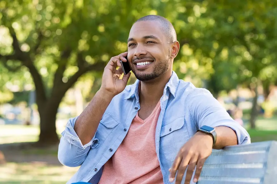 Smiling man talking on phone while sitting on a park bench outdoors among green trees.