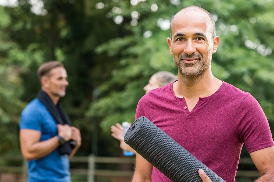Smiling man holding a yoga mat outdoors with others nearby, preparing for a holistic wellness session.