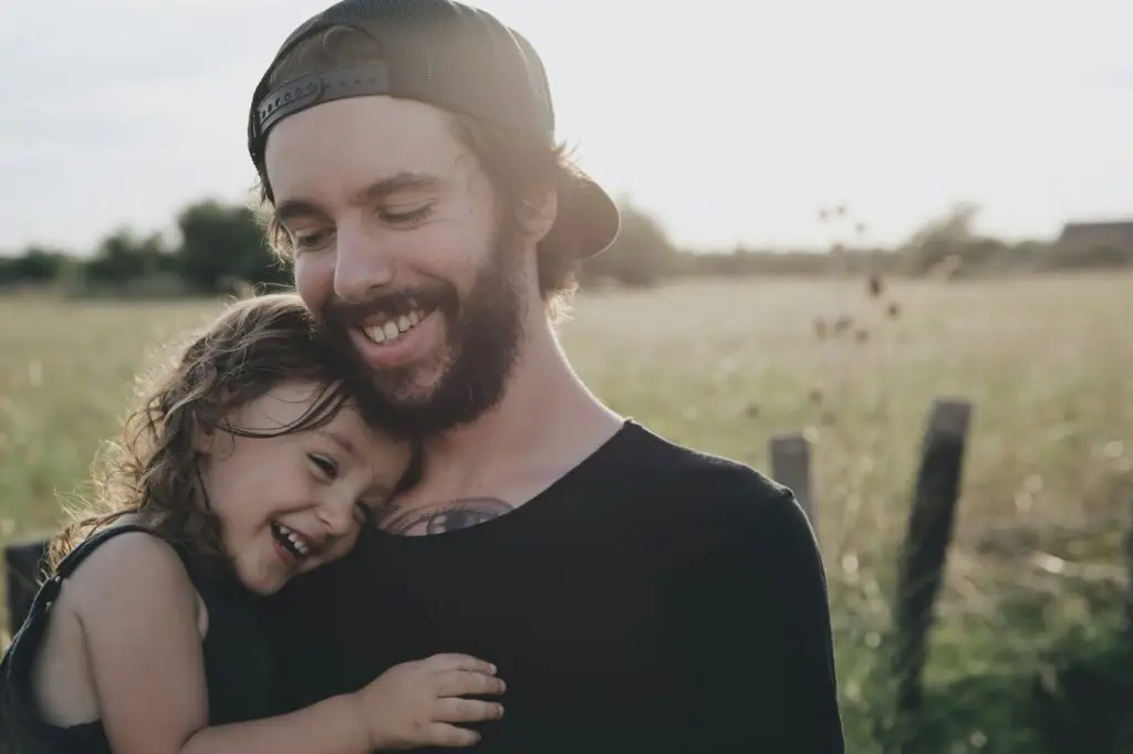 Smiling father holding his laughing young daughter in a sunlit field, sharing a joyful embrace outdoors.