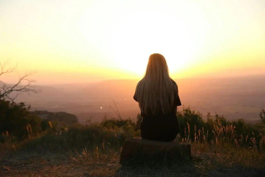 Woman sitting alone on a hilltop watching the sunset over a valley, reflecting in peaceful solitude.