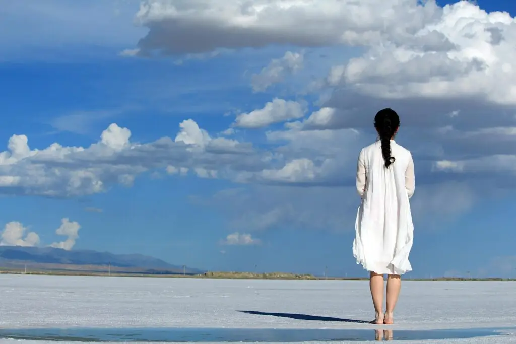 Woman in white dress standing alone facing open horizon under a vast blue sky with clouds, symbolizing hope and recovery.
