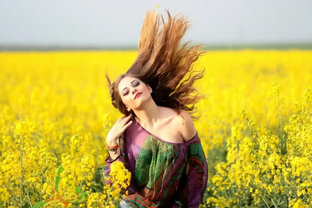 Woman with flowing hair standing peacefully in a bright yellow flower field under an open sky.