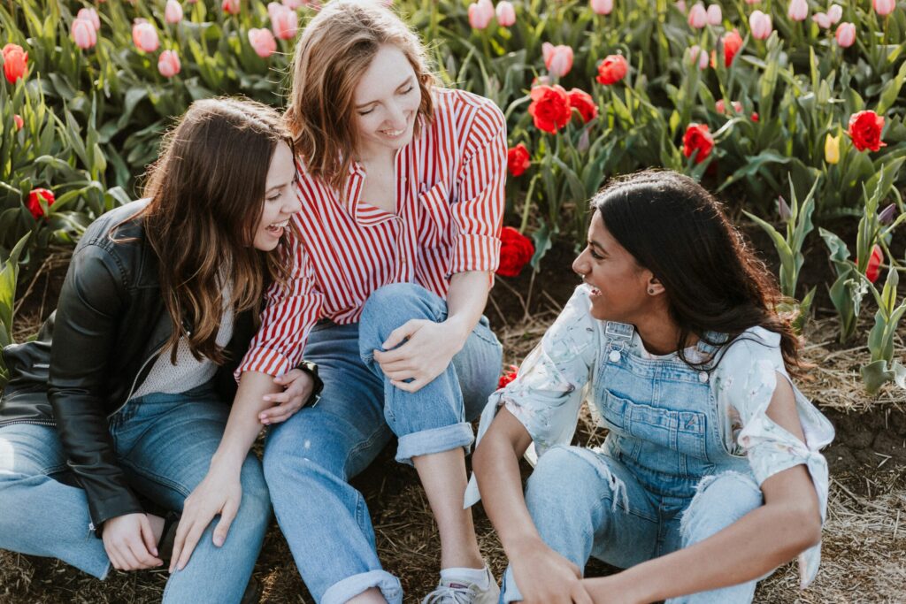 Three women sitting together and laughing in a field of pink and red tulips, sharing a supportive moment.