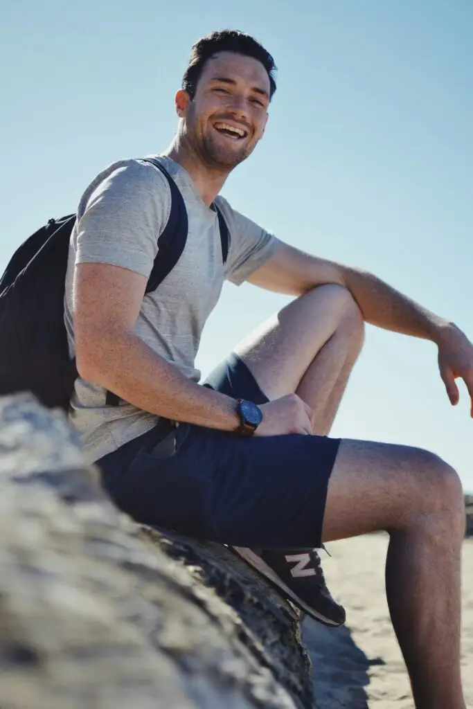 Smiling young man with a backpack sitting on a rock outdoors under a clear blue sky.