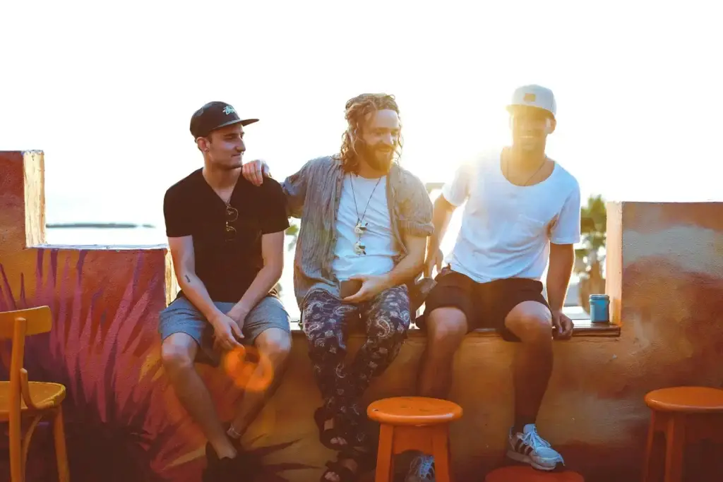 Three young men sitting together outdoors at sunset, talking and smiling in a relaxed, supportive moment.