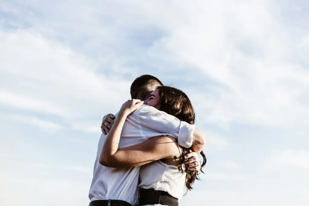 A man and woman embrace in a tight, emotional hug against a bright, cloudy sky.
